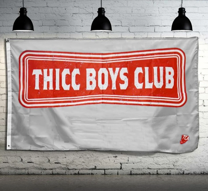 The Team Thicc Boys Club Flag by Tango Charlie Apparel, with a red rectangular THICC BOYS CLUB emblem on white polyester, hangs on a white brick wall. Black industrial lights highlight its double-stitching and metal grommets, while a TCA logo appears in the bottom right corner.