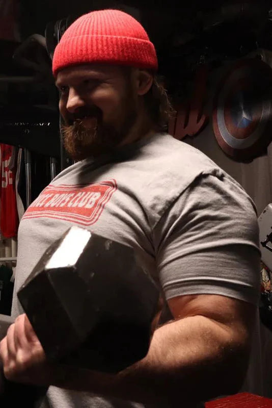 A bearded man in a red beanie lifts a large dumbbell in the gym, smiling. He wears a gray THICC Boys Club T-Shirt by Tango Charlie Apparel, with weights and plates energizing the background.