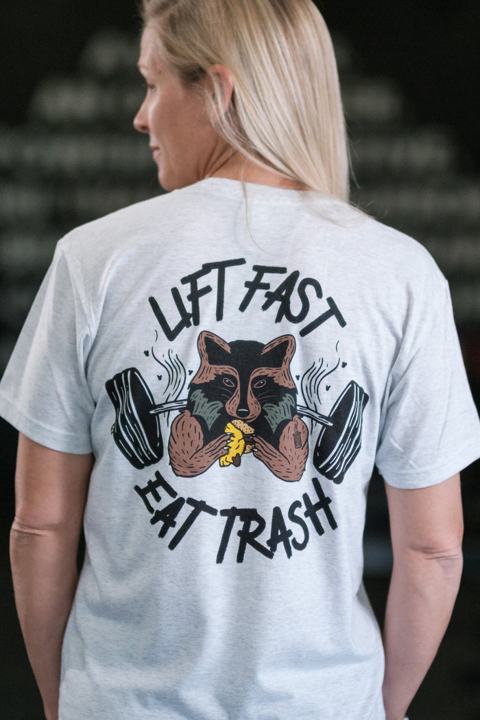 Wearing a tri-blend gray Trash Panda Weight Lifting Club tee from Tango Charlie Apparel, a person showcases a trash panda lifting weights and munching on a banana, with the text Lift Fast, Eat Trash.