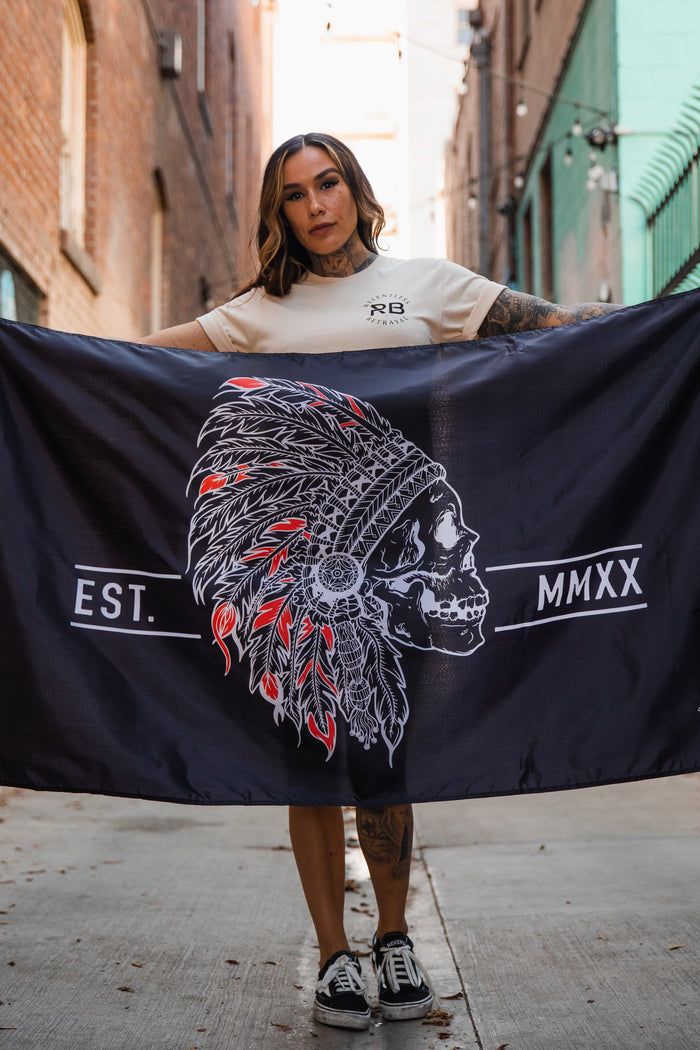 In an alleyway with brick and teal walls, a person in a beige t-shirt and black sneakers holds the Trustless Chief Flag by Relentless Betrayal, featuring a skull in a feathered headdress labeled EST. MMXX. Made of durable polyester with metal grommets for longevity.