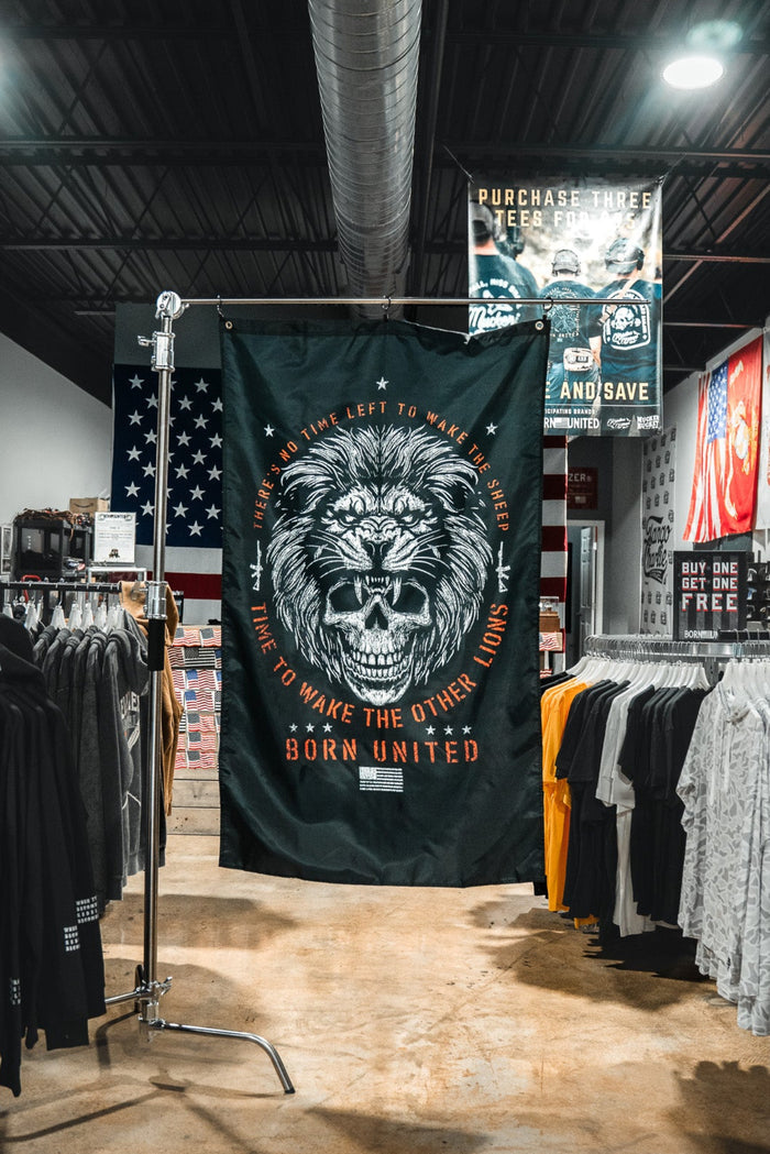 A clothing store showcases the Born United Private Label Waking Lions Flag, featuring a roaring lion and the phrase “It takes one left to wake the others. Born United,” displayed among shirts and hoodies under bright lights.