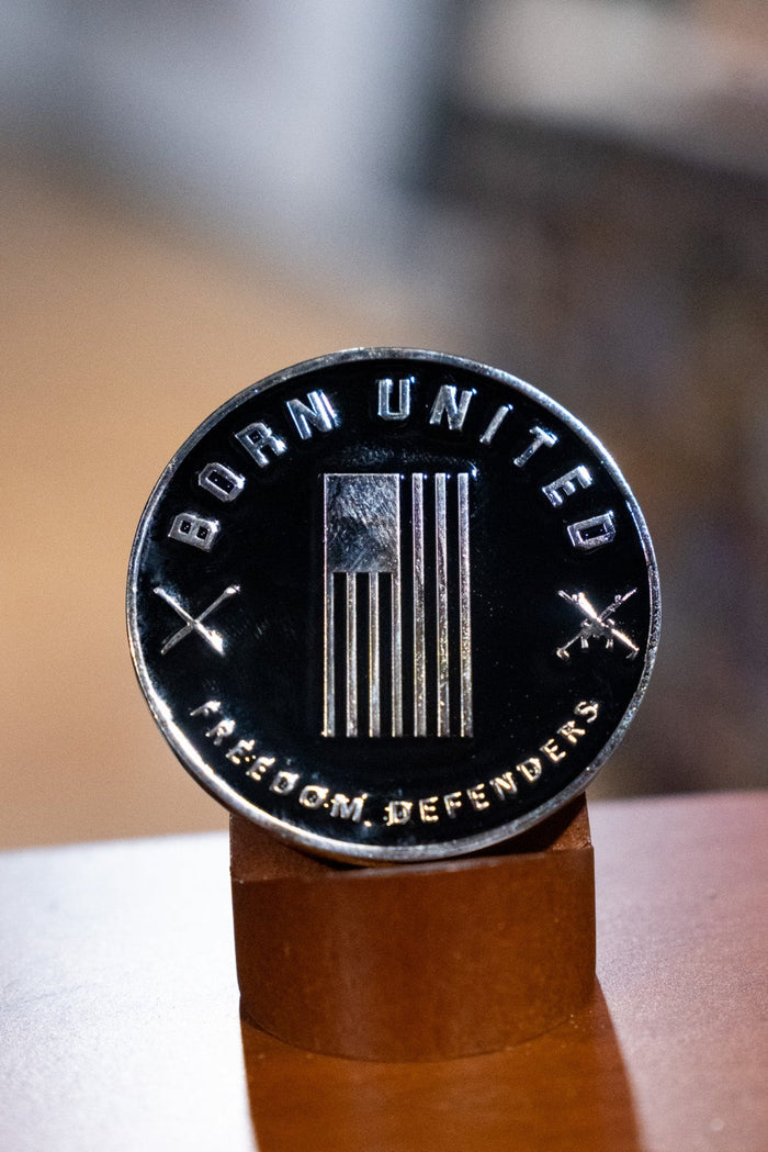 The Warrior Spirit Challenge Coin by Born United features a stylized American flag, โBORN UNITEDโ above, โFREEDOM DEFENDERSโ below, and crossed riflesโsymbolizing true warrior spiritโdisplayed on a wooden stand.