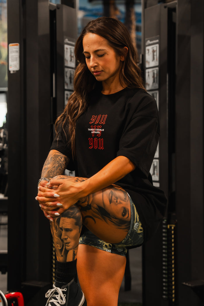 A woman with long brown hair stretches her leg in a gym, wearing the Tango Charlie Apparel You vs You - Premium Oversized Tee and shorts that reveal portrait tattoos on her leg. Fitness equipment is visible in the background.