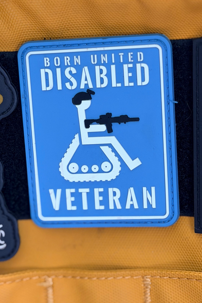 The Born United Private Label Disabled Vet PVC Patch features a blue design with a stylized wheelchair user holding a rifle. It’s attached to yellow fabric and displays the text “BORN UNITED DISABLED VETERAN.”.