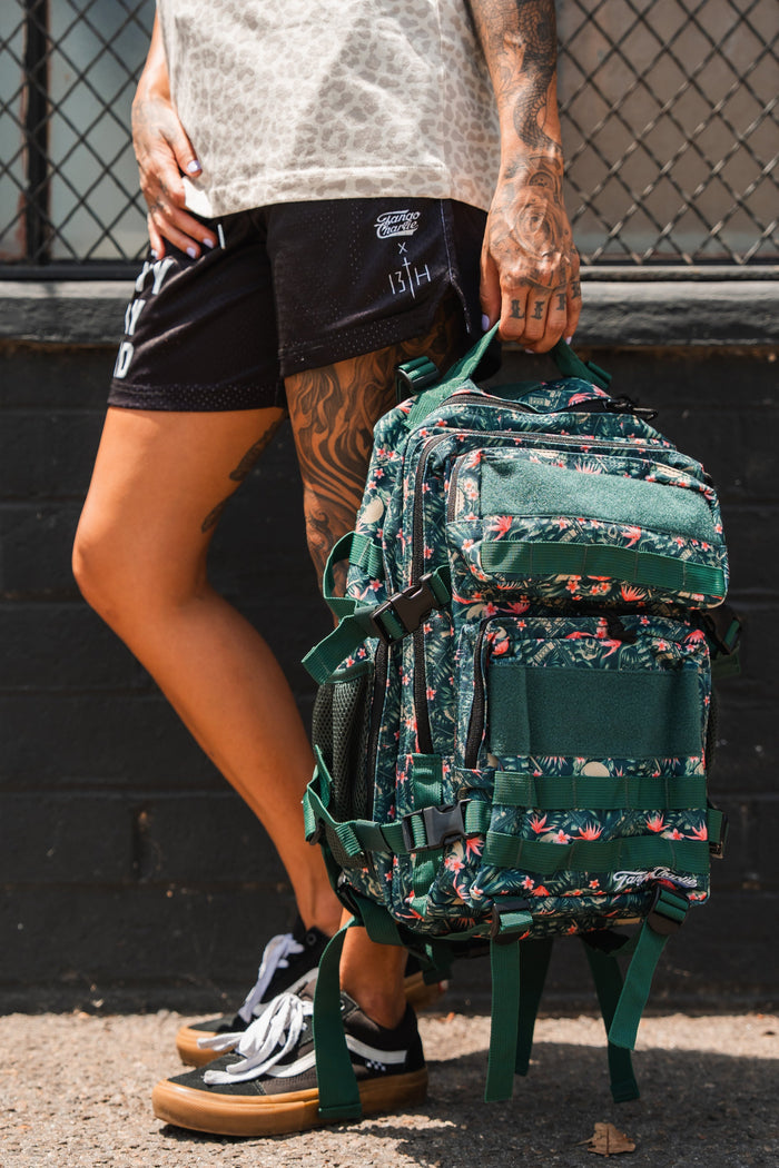 A tattooed individual stands on a sidewalk holding the Aloha AR - Tac Pac 25L by Tango Charlie Apparel, a green, floral-patterned waterproof backpack. They're dressed in black shorts, a patterned shirt, and black sneakers; a wire fence and brick wall are behind them.