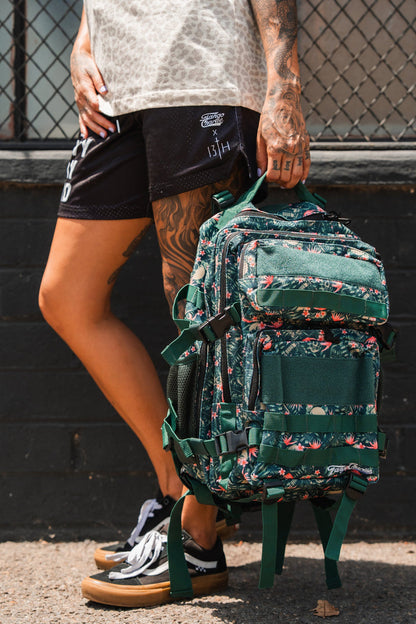 A tattooed individual stands on a sidewalk holding the Aloha AR - Tac Pac 25L by Tango Charlie Apparel, a green, floral-patterned waterproof backpack. They're dressed in black shorts, a patterned shirt, and black sneakers; a wire fence and brick wall are behind them.