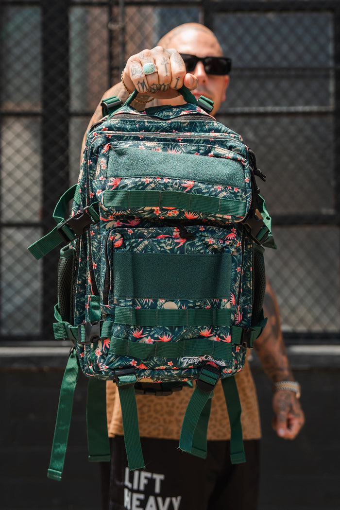 A tattooed person in sunglasses holds up a Tango Charlie Apparel Aloha AR - Tac Pac 25L waterproof floral backpack, hiding their face outdoors by a metal fence and window.