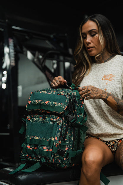 A woman with long hair and tattoos sits indoors, holding the Tango Charlie Apparel Aloha AR - Tac Pac 25L backpack with a green floral pattern on her lap. She wears a beige shirt and shorts in a dimly lit gym setting.