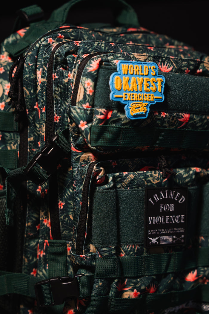 The image shows the sharply focused upper half of the Tango Charlie Apparel Aloha AR - Tac Pac 25L, a floral-patterned waterproof backpack with a blue “World’s Okayest Exerciser” patch and a black “Trained for Violence” patch.