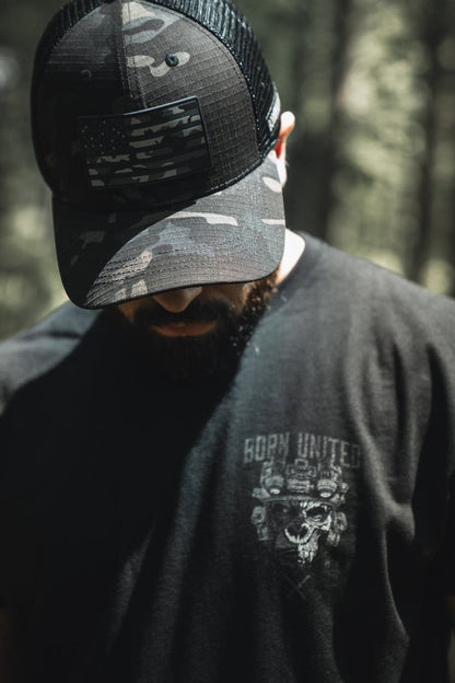 A person in a Born United Private Label Born United Black Camo Trucker - Camo Flag hat and a black t-shirt stands outdoors with head bowed, forest background blurred.