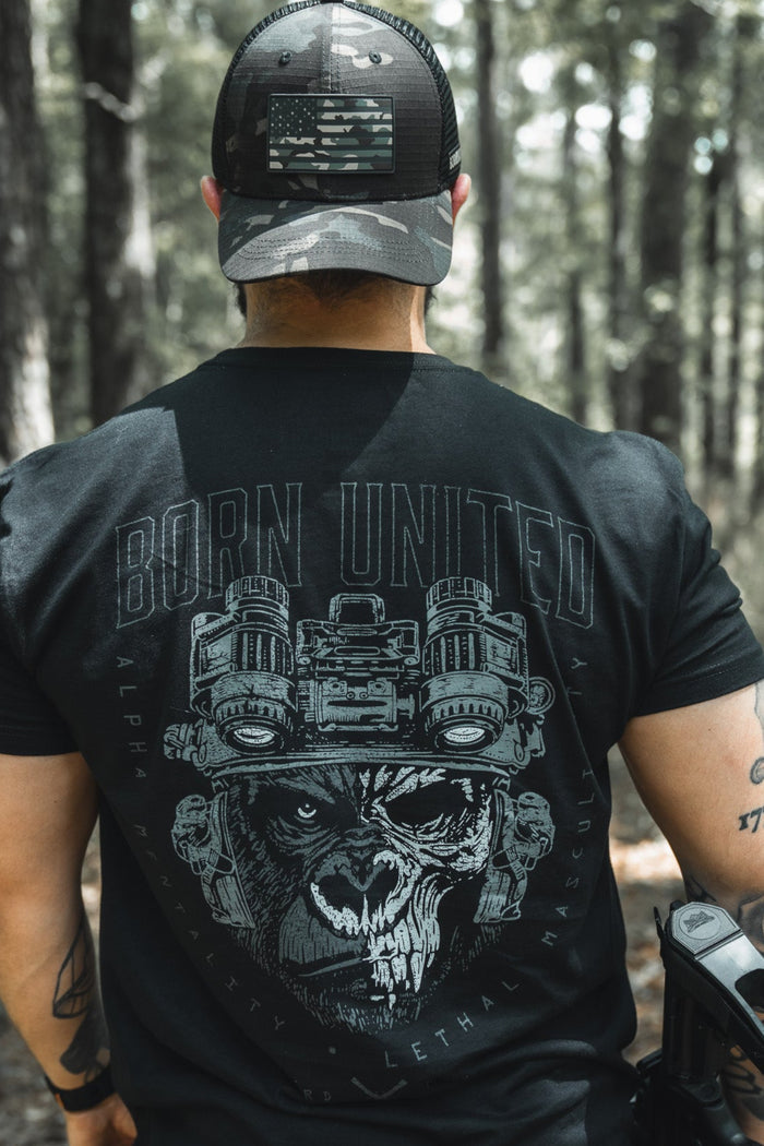 A man stands in a sunlit forest wearing a black Born United shirt with a wolf graphic, the Born United Black Camo Trucker – Camo Flag hat, and carrying a holstered gun. Trees and sunlight fill the background. Brand: Born United Private Label.