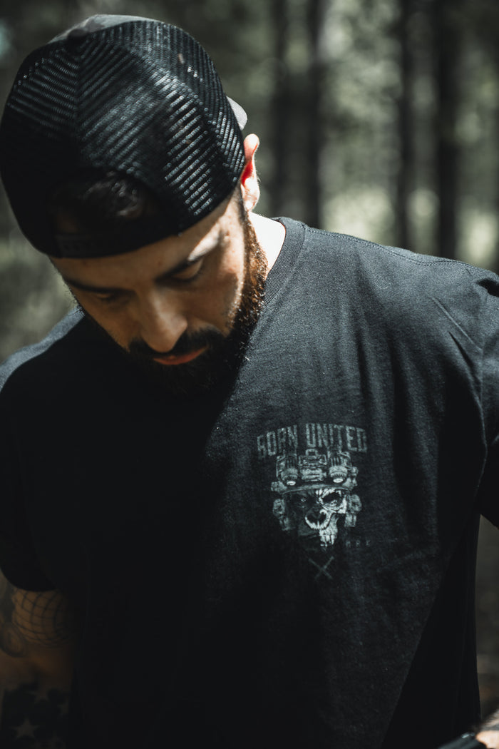A man in a Born United Black Camo Trucker - Camo Flag hat by Born United Private Label and a black T-shirt with a skull and Born United graphic looks down in a sunlit wooded area.