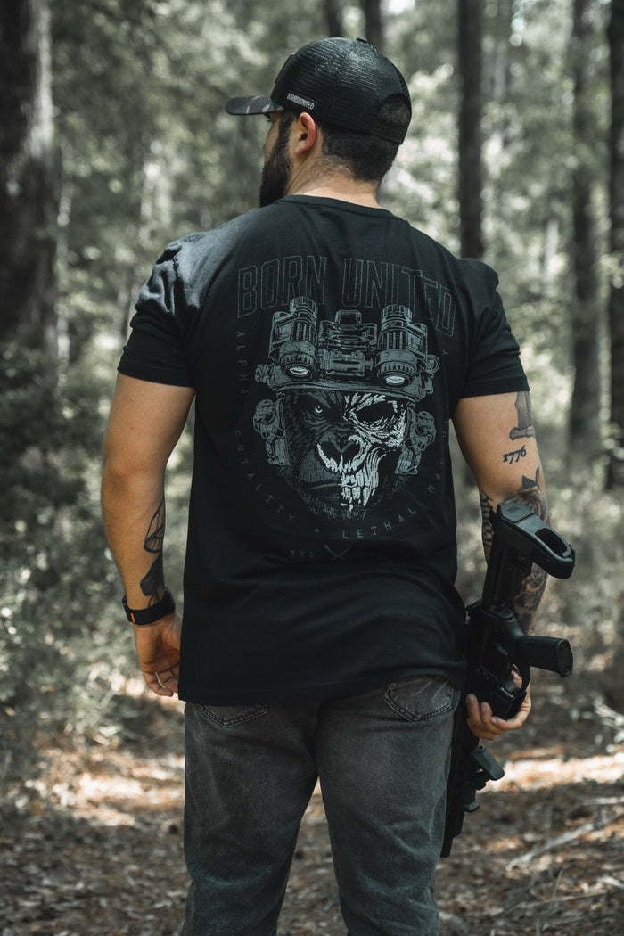 A man stands in a wooded area, wearing a black cap and the Alpha Mentality tee from Born United Private Label, featuring a striking skull graphic on the back. Sunlight filters through the trees, highlighting this bold men's shirt.