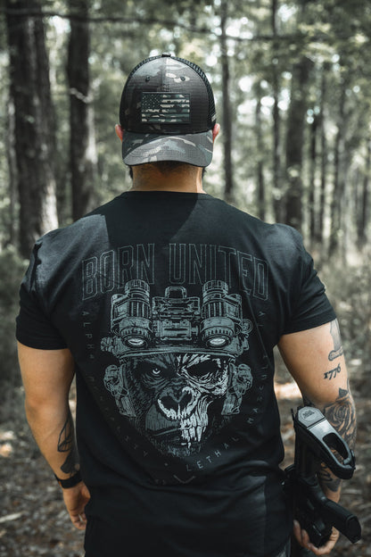 A man stands in a forest wearing a black cap with a U.S. flag patch and the Born United Private Label "Alpha Mentality" men's tee, featuring a skull with night vision goggles. He has tattoos on his left arm and a holster on his right hip.