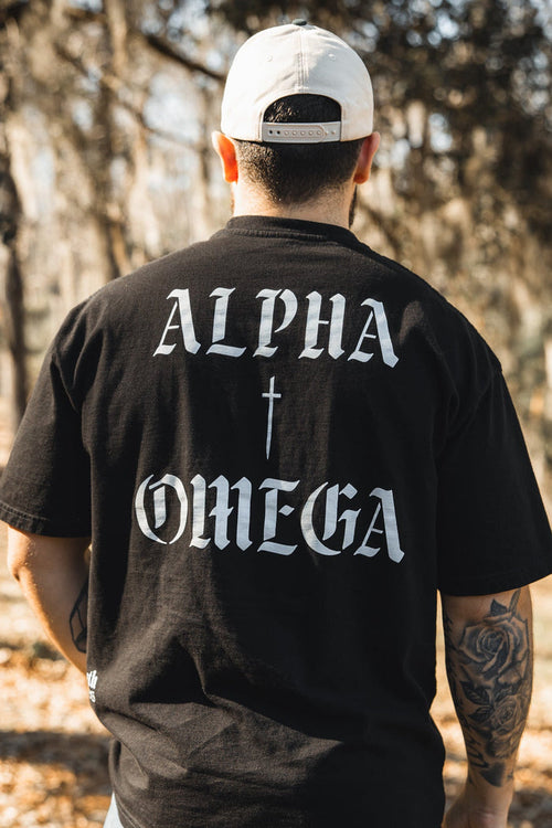 A man stands outdoors with his back to the camera, wearing the 13th Alpha & Omega Premium Oversized Tee - Black, featuring bold white "ALPHA" and "OMEGA" text with a cross—ideal for Christian streetwear enthusiasts.