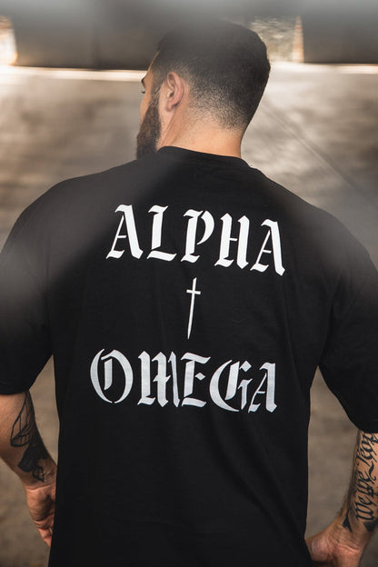 A man with short hair and a beard faces away, wearing the 13th Alpha & Omega Premium Oversized Tee in black—an oversized heavyweight cotton shirt with bold white gothic “ALPHA” and “OMEGA,” a distinctive piece of Christian streetwear.