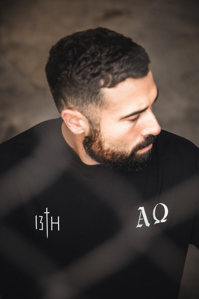 A bearded man with short dark hair looks down, wearing the 13th Alpha & Omega Premium Oversized Tee in black, featuring bold white symbols. The blurred outdoor background highlights the premium heavyweight cotton streetwear.