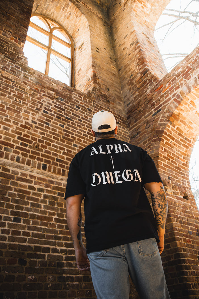 A person wears the 13th Alpha & Omega Premium Oversized Tee in black and a white cap, standing in a brick building with arched windows, looking upward as sunlight streams through a large overhead window.