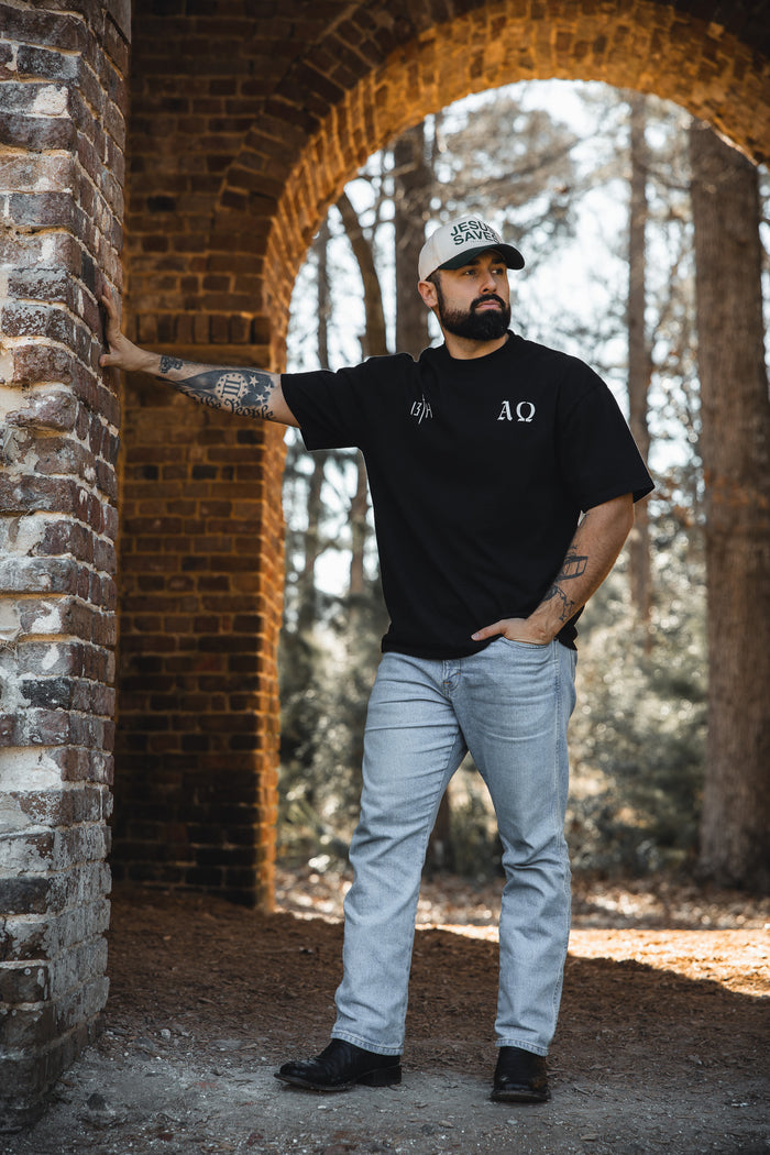 A bearded man in the 13th Alpha & Omega Premium Oversized Tee in black, light cap, light blue jeans, and black shoes leans on a brick archway outdoors with trees and sunlight in the background.