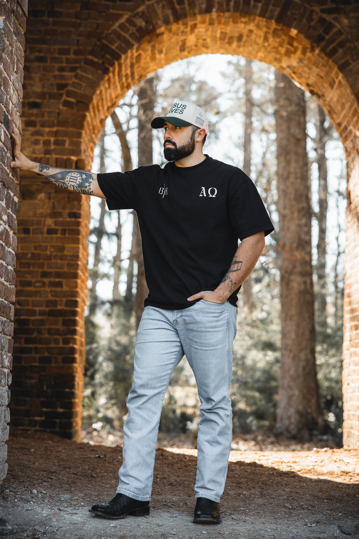 A tattooed, bearded man stands under a brick archway wearing the 13th Alpha & Omega Premium Oversized Tee in black, light blue jeans, black shoes, and a baseball cap. One hand rests on the wall, with trees visible in the background.