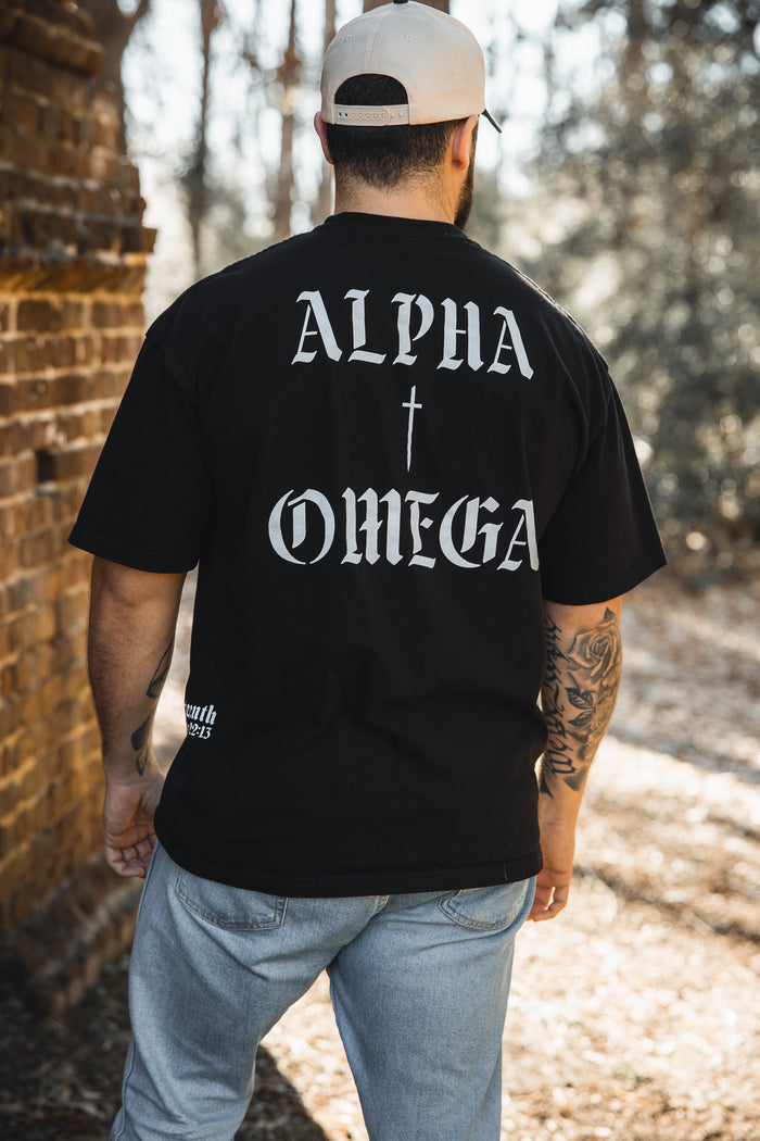 A man in a beige cap and the 13th Alpha & Omega Premium Oversized Tee - Black stands outdoors by a brick wall, highlighting bold Christian streetwear amidst trees and sunlight.