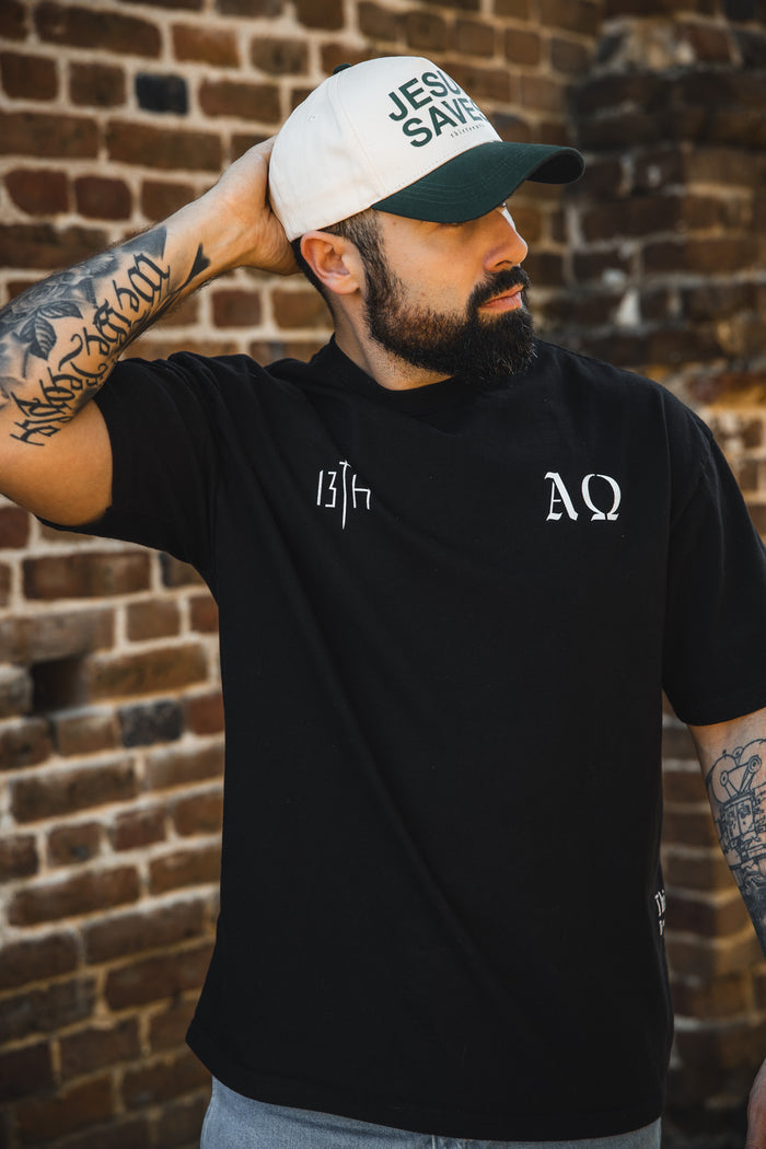 A tattooed man stands in front of a brick wall, looking sideways with one hand behind his head, wearing a black 13th Alpha & Omega Premium Oversized Tee with white Greek letters and a white JESUS SAVES cap.