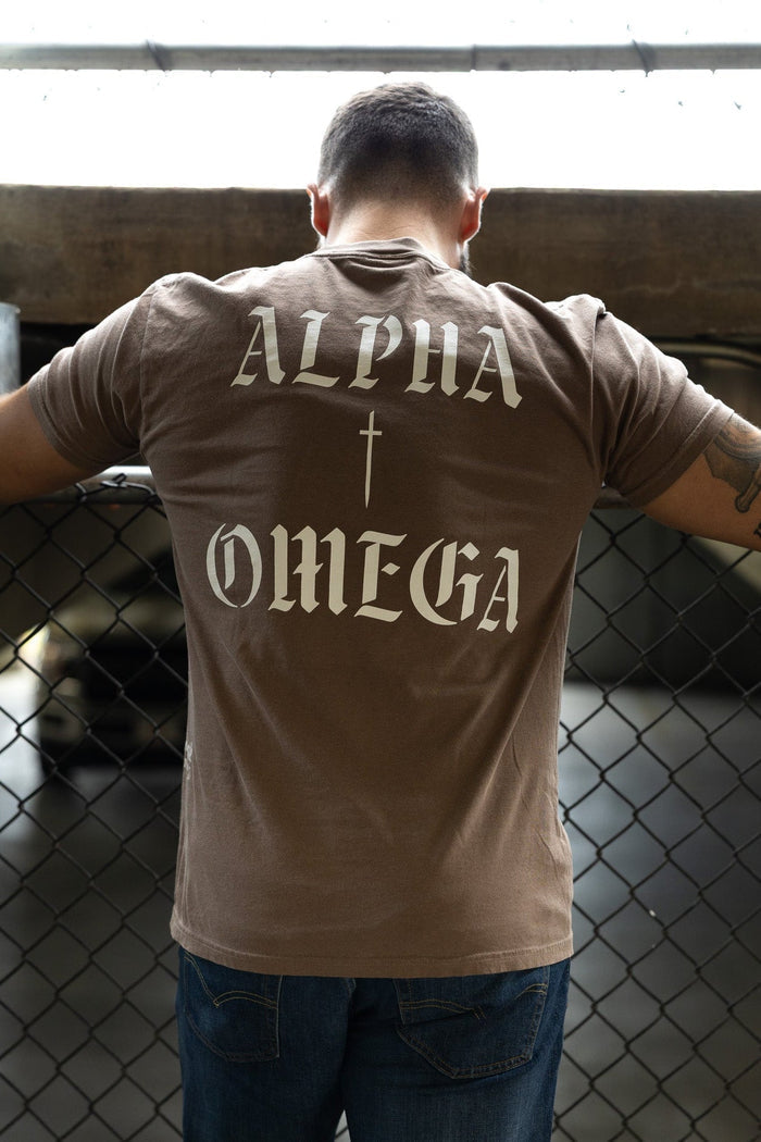 A man leans against a chain-link fence wearing the 13th Alpha & Omega Premium Oversized Tee in Espresso, featuring bold white "Alpha & Omega" lettering and a cross on the back.