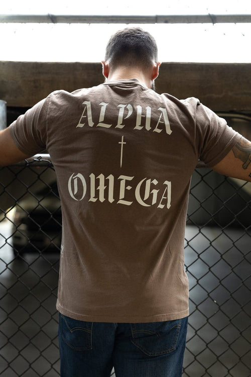 A man leans against a chain-link fence wearing the 13th Alpha & Omega Premium Oversized Tee in Espresso, featuring bold white "Alpha & Omega" lettering and a cross on the back.