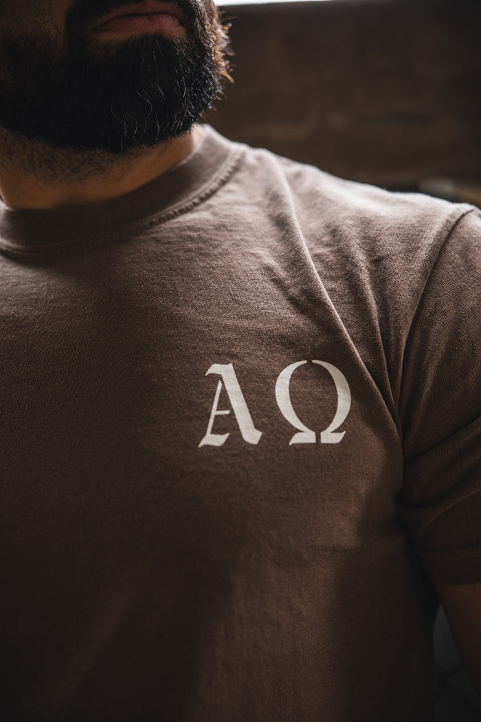 A bearded person wears the 13th Alpha & Omega Premium Oversized Tee in Espresso, showcasing white ΑΩ symbols on the chest. The image highlights the shirt's unique design by focusing on the upper torso.