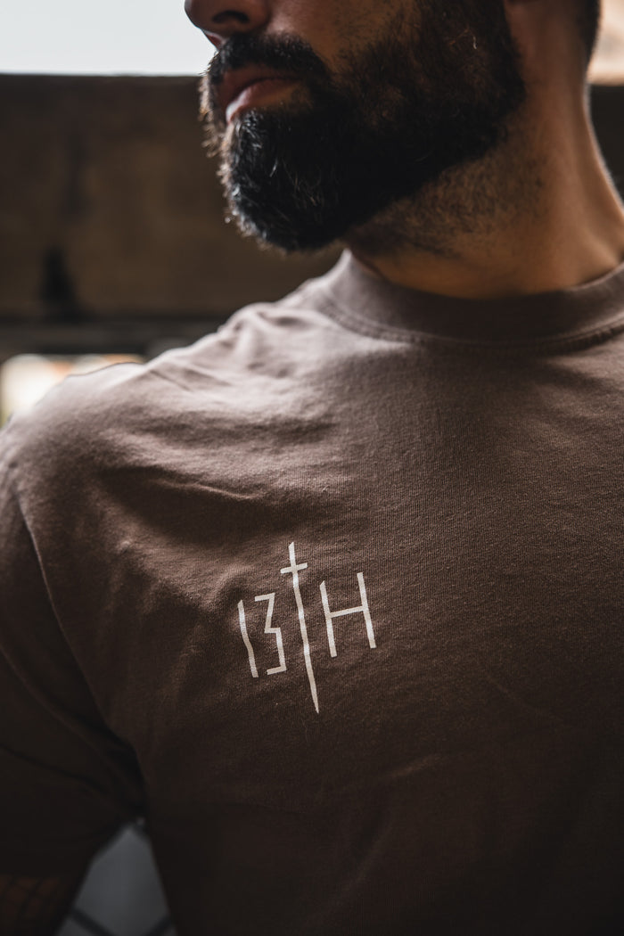 A bearded man wears the 13th Alpha & Omega Premium Oversized Tee in Espresso, featuring a white chest design with the number 13, a sword, and an H. The photo is a shoulders-up shot.