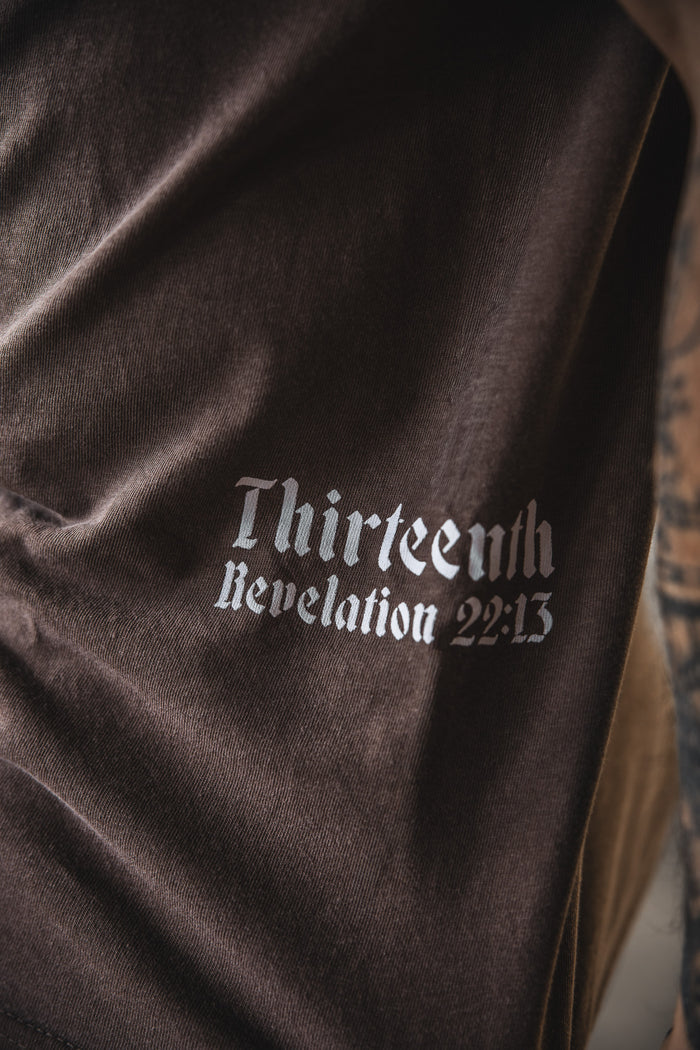 Close-up of the 13th Alpha & Omega Premium Oversized Tee in Espresso, featuring white Gothic "Thirteenth Revelation 22:13" text. The shirt's brown fabric and partial tattoos on a nearby arm are visible.