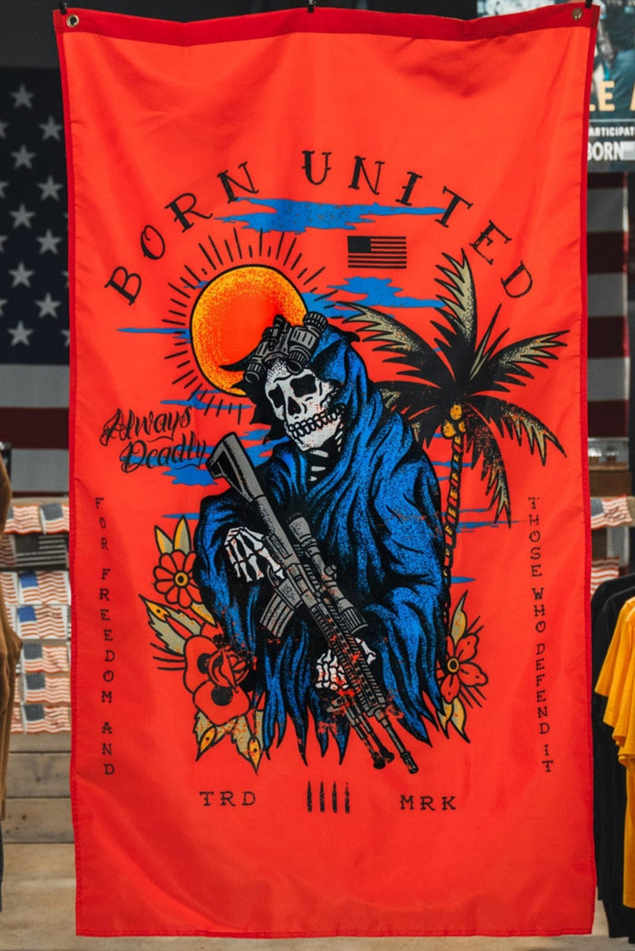 A red Always Deadly Flag by Born United Private Label, featuring a colorful skeleton in a blue cloak with a rifle, palm trees, and a setting sun, hangs in the clothing store. The banner reads "Born United" and "Anatomy of Peaceful.
