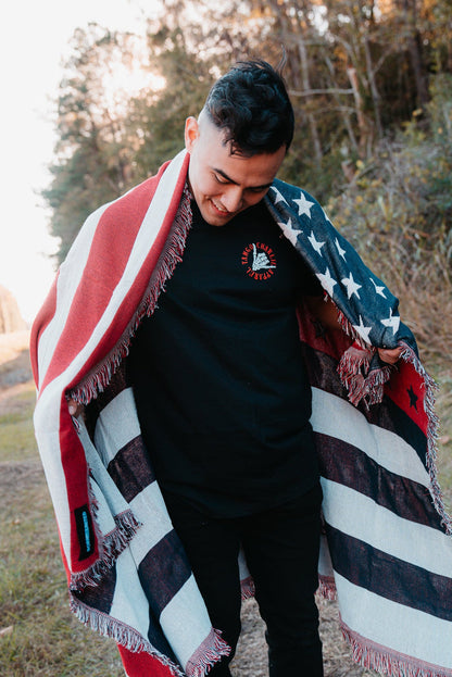 The person stands outdoors, wrapped in the Born United Private Labels American Flag Jacquard Blanket, smiling as they look down. They are dressed in a black t-shirt and pants, with trees and a clear sky in the background.