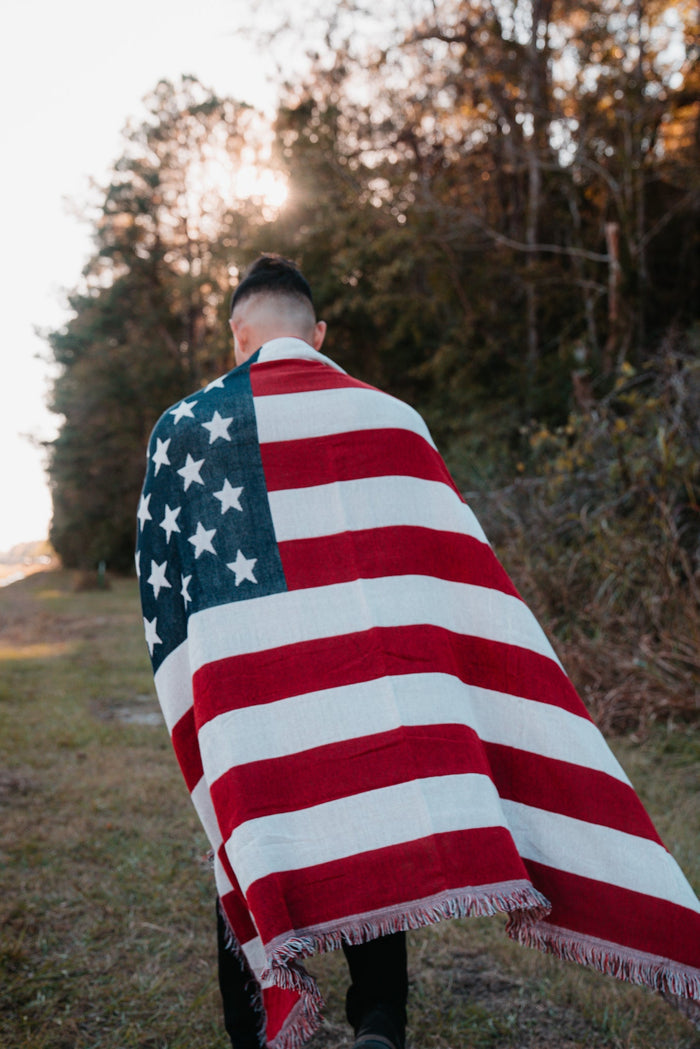 Wrapped in the American Flag Jacquard Blanket by Born United Private Label, a person strolls along a grassy path. The setting sun casts a warm light through the trees, creating a serene backdrop for the journey.