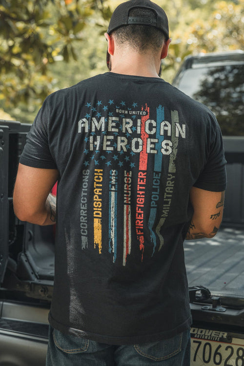 A man stands at the back of a pickup truck, wearing a black cap and an American Heroes Tee by Born United Private Label, featuring colorful stripes and words like Corrections, Dispatch, Firefighter, Police, and Military.