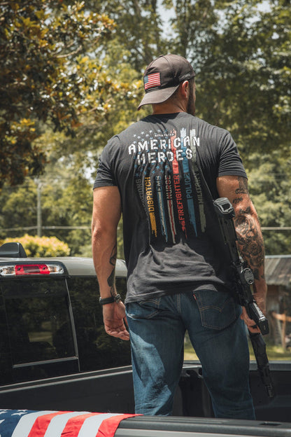 A man stands in the back of a pickup, wearing a Born United Private Label American Heroes Tee with colored lines and names on the back. He holds a rifle, has an American flag patch on his cap, and an American Heroes Flag draped nearby.