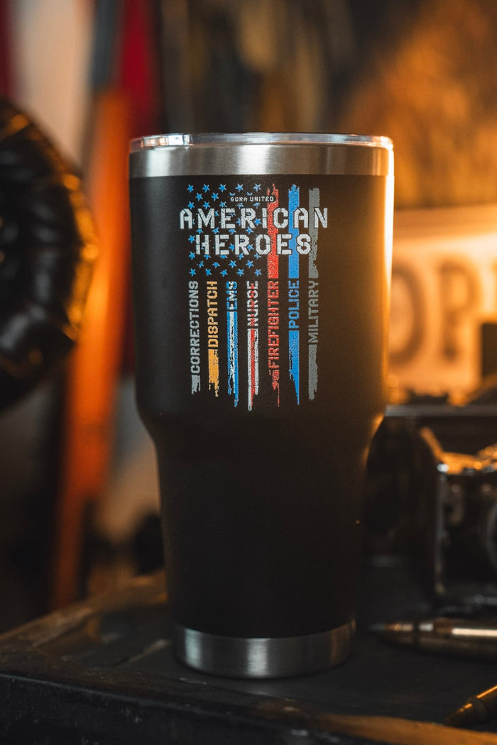 The 30oz American Heroes Tumbler by Born United Private Label, featuring colored lines for corrections, dispatch, EMS, fire, nurses, police & military, sits on a dark surface with blurred background items and a sign.