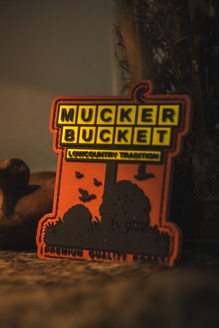 An orange and black 'Mucker Bucket' outdoor sign with a silhouette of a campfire, trees, and birds, set against a dark, moody background with a duck toy on the left.