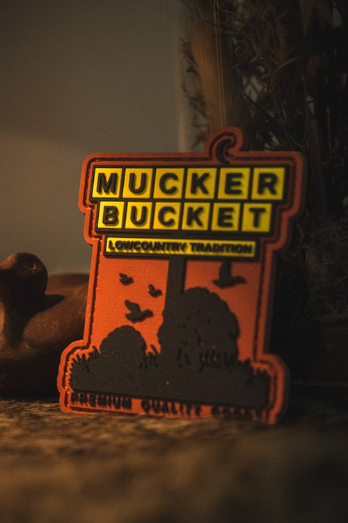 An orange and black 'Mucker Bucket' outdoor sign with a silhouette of a campfire, trees, and birds, set against a dark, moody background with a duck toy on the left.