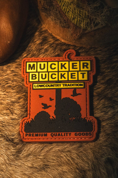 A rectangular embroidered patch with a rustic outdoor design, featuring the text 'MUCKER BUCKET' and 'LOWCOUNTRY TRADITION' on a brown fur background.