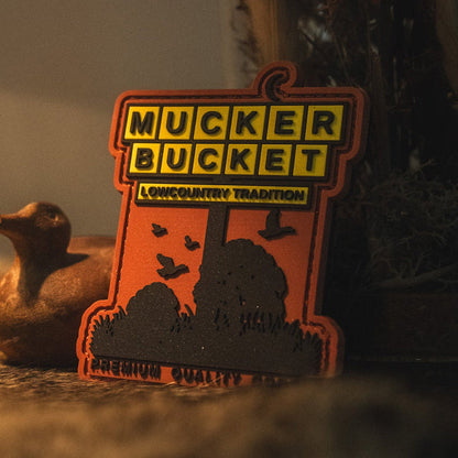A sign reading Mucker Bucket Lowcountry Tradition Premium Quality sits on a surface next to a small bird figurine and the American Tradition PVC Patch by Mucker Bucket, with plants in the background.