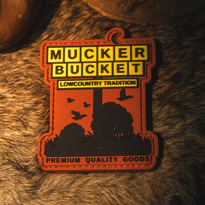 The American Tradition PVC Patch by Mucker Bucket features "MUCKER BUCKET," "LOWCOUNTRY TRADITION," and "PREMIUM QUALITY GOODS" text, along with a detailed silhouette of a tree, birds, and bushes, displayed on a textured surface. This 4x4 patch is expertly crafted from durable 3D PVC and equipped with Velcro backing for easy attachment.