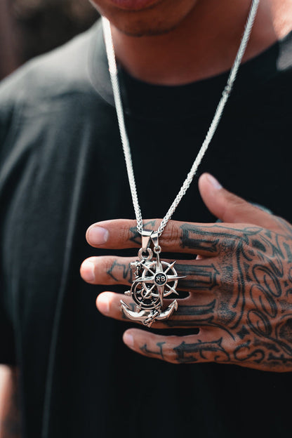 A person with tattooed fingers holds the Anchors Aweigh Necklace by Relentless Betrayal. Its handcrafted silver pendant features nautical symbols, elegantly hanging from a stainless steel chain, ensuring both style and a lifetime warranty—all set against a black shirt.