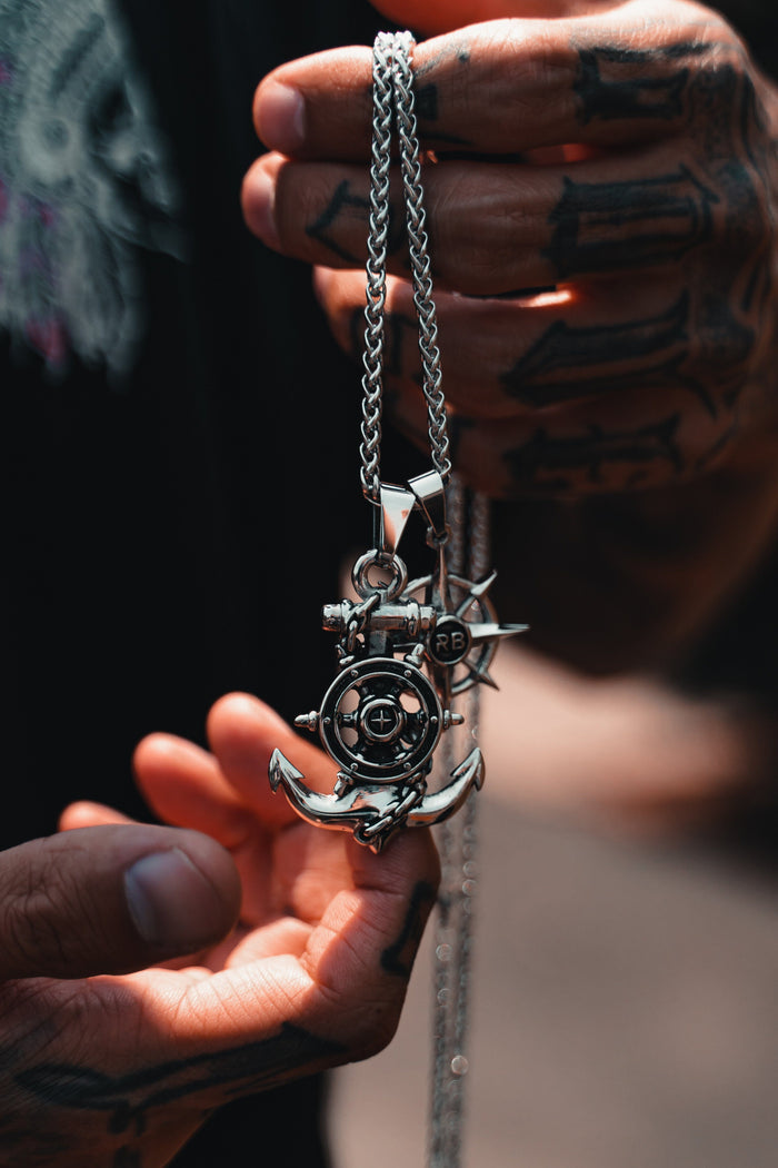 A person with tattooed hands showcases the Anchors Aweigh Necklace by Relentless Betrayal, a handcrafted silver pendant featuring an anchor and ships wheel. Hanging from a stainless steel chain, it stands out against a blurred background, exuding timeless elegance.