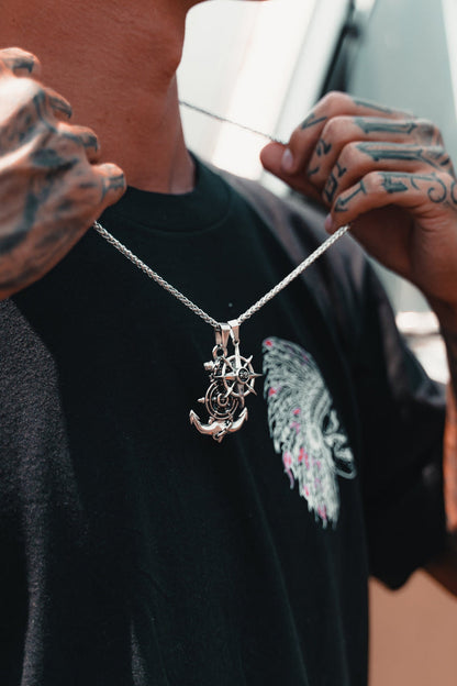 A tattooed individual in a black t-shirt with wings displays the Anchors Aweigh Necklace from Relentless Betrayal, featuring a handcrafted silver crescent moon, star, and anchor on a stainless steel chain, while outdoors in sunny weather.