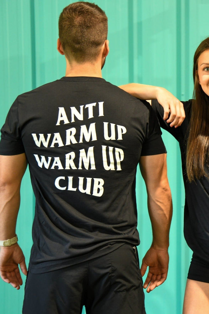 In a gym with teal walls and orange equipment, a man with his back to the camera and a smiling woman face forward, both wearing Tango Charlie Apparels black Anti Warmup, Warmup Club T-Shirts, creating an anti-social vibe.
