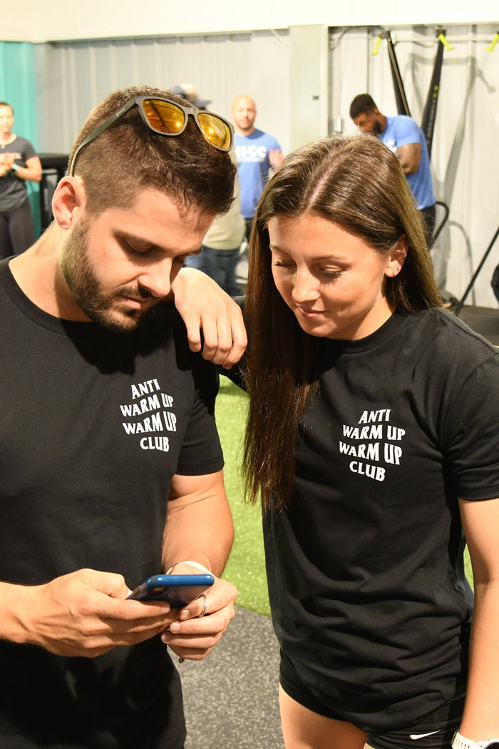 Two people in matching Tango Charlie Apparel Anti Warmup, Warmup Club black T-shirts stand in a gym, exuding an anti-social vibe. The man checks his phone as the woman leans in to see, both ignoring the buzz of workout equipment and fellow gym-goers.