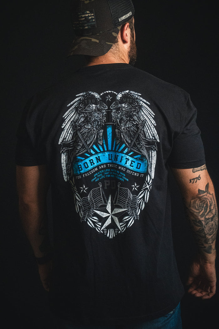 A man is wearing a black Born United Private Label "Back the Badge" cap and a matching black T-shirt featuring an eagle, wings, stars, and the phrases "Born United" and "For freedom and those who defend it" on the back. A tattoo is visible on his left arm.