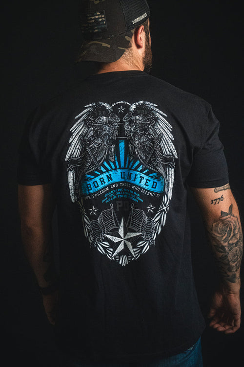 A man is wearing a black Born United Private Label "Back the Badge" cap and a matching black T-shirt featuring an eagle, wings, stars, and the phrases "Born United" and "For freedom and those who defend it" on the back. A tattoo is visible on his left arm.