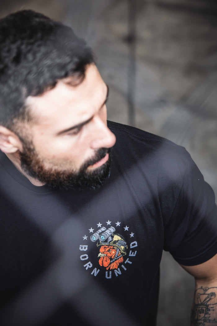A bearded man with short dark hair wears a black Battle Bird t-shirt by Born United Private Label, made from a 60/40 blend and featuring a heart, camera, and stars graphic. The background is neutral and out of focus.